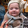 baby, child, smile, hat, coat, hand, red_nail_polish, plaid, fuzzy, indoor, person, blurred_background, face, cute, warm_clothing, portrait, holding, infant, happy, cozy