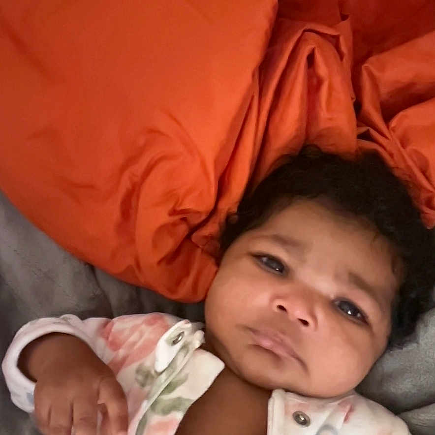 Mylanii is registered to the contest to win money with this photo: baby, bed, blanket, child, clothing, curious_expression, cute, dark_hair, face, floral_onesie, hand, indoor, infant, lying_down, newborn, orange_blanket, person, portrait, skin, sleepwear