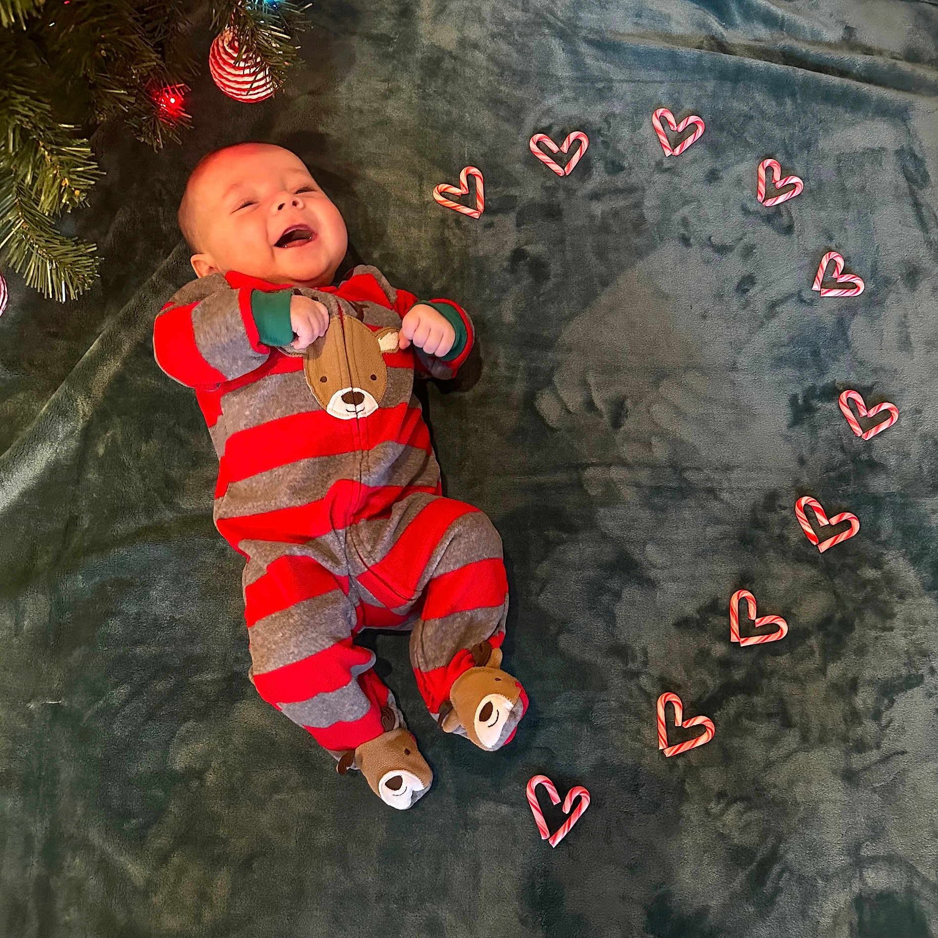 Caspian is registered to the contest to win money with this photo: baby, bear_slippers, blanket, candy_canes, child, christmas_tree, cute, festive, gray, green, happy, hearts, holiday, indoors, infant, lying_down, red, smiling, soft_texture, striped_onesie