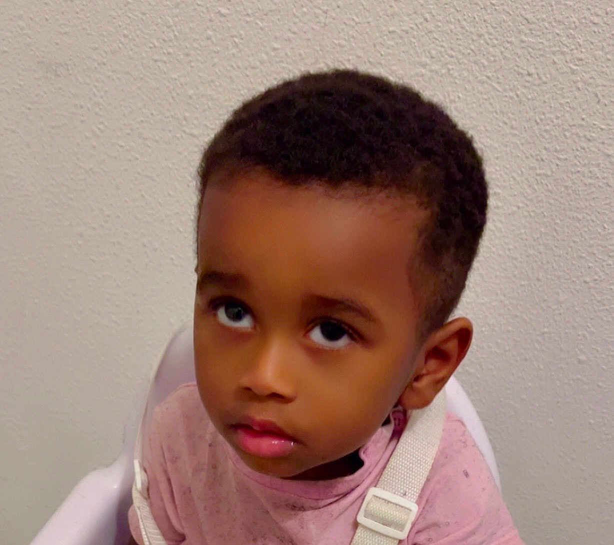 Ilyan participe au concours pour gagner de l'argent avec cette photo : child, toddler, curious, face, eyes, high_chair, pink_shirt, white_strap, indoor, portrait, young_child, looking_up, wall, expression, person, human, sitting, short_hair, skin, casual_clothing