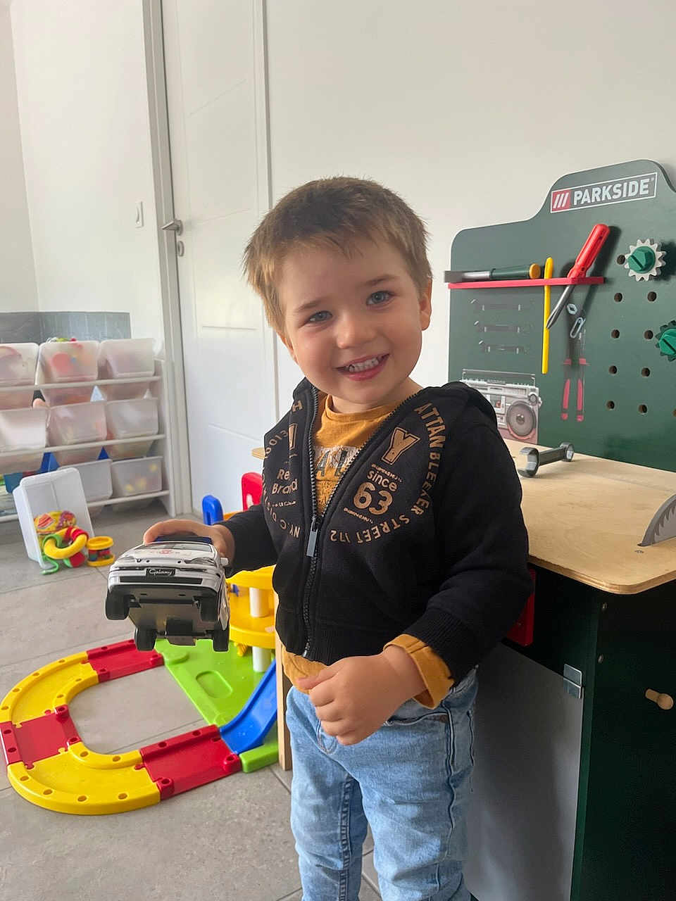 Gabriel participe au concours pour gagner de l'argent avec cette photo : child, toy_car, indoor, smiling, playroom, toy_workbench, colorful, jeans, hoodie, floor, plastic_toys, storage_bins, door, person, young_child, happy, playing, furniture, casual_clothing, blue_eyes