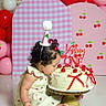 balloons, birthday, birthday_cake, cake, cake_stand, celebration, cherries, child, curly_hair, dessert, dress, festive, indoors, party_hat, pink, portrait, red, sitting, toddler, white