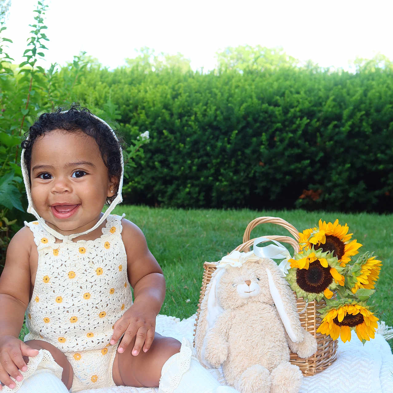 Parker is registered to the contest to win money with this photo: baby, smiling, outdoor, grass, blanket, sunflowers, basket, plush_toy, bunny, cute, child, greenery, garden, happy, portrait, clothing, headwear, sitting, sock, nature