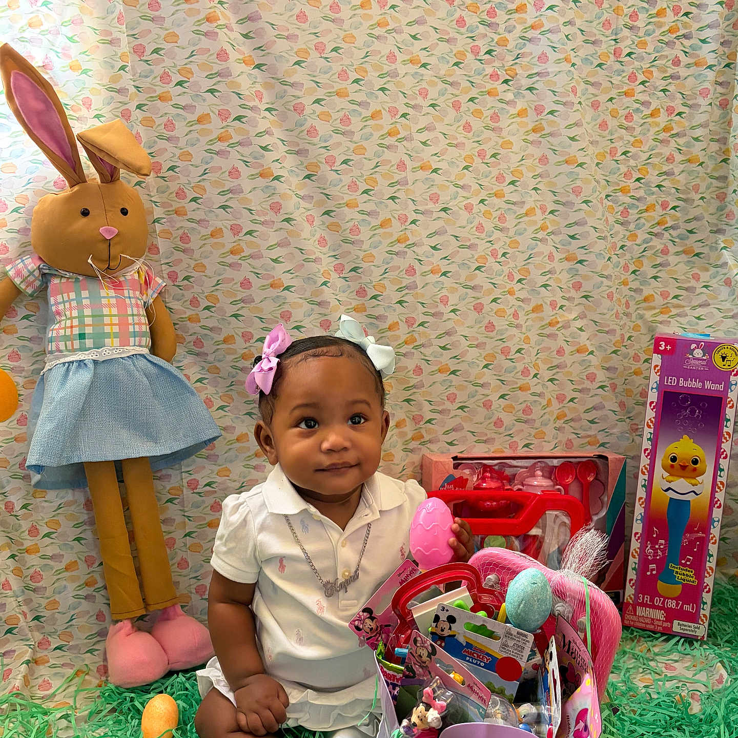 Kapri is registered to the contest to win money with this photo: toddler, child, toy, stuffed_animal, easter, basket, grass, bow, white_clothing, indoor, cute, smiling, holiday, pink, blue, gift, celebration, easter_egg, sitting, portrait