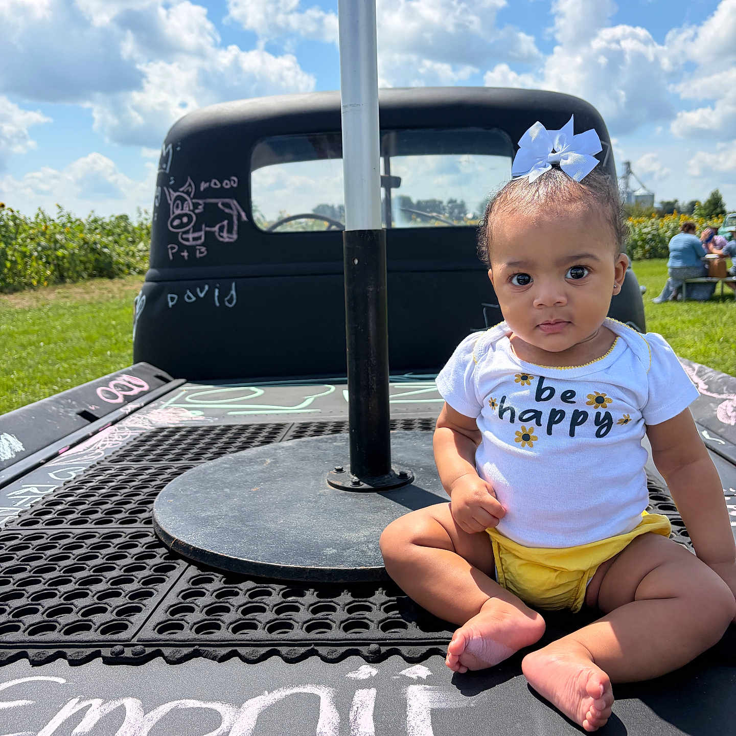 Emonii is registered to the contest to win money with this photo: baby, child, outdoor, sky, clouds, vehicle, chalk, writing, bow, shirt, yellow_clothing, grass, sunny, portrait, face, sitting, cute, expression, people_in_background, field