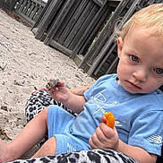 Bodie joined the competition — help win amazing prizes! toddler, child, outdoor, playground, sand, wooden_structure, fence, blue_clothing, snack, food, sitting, person, face, curious, daylight, nature, play, casual, cute, portrait