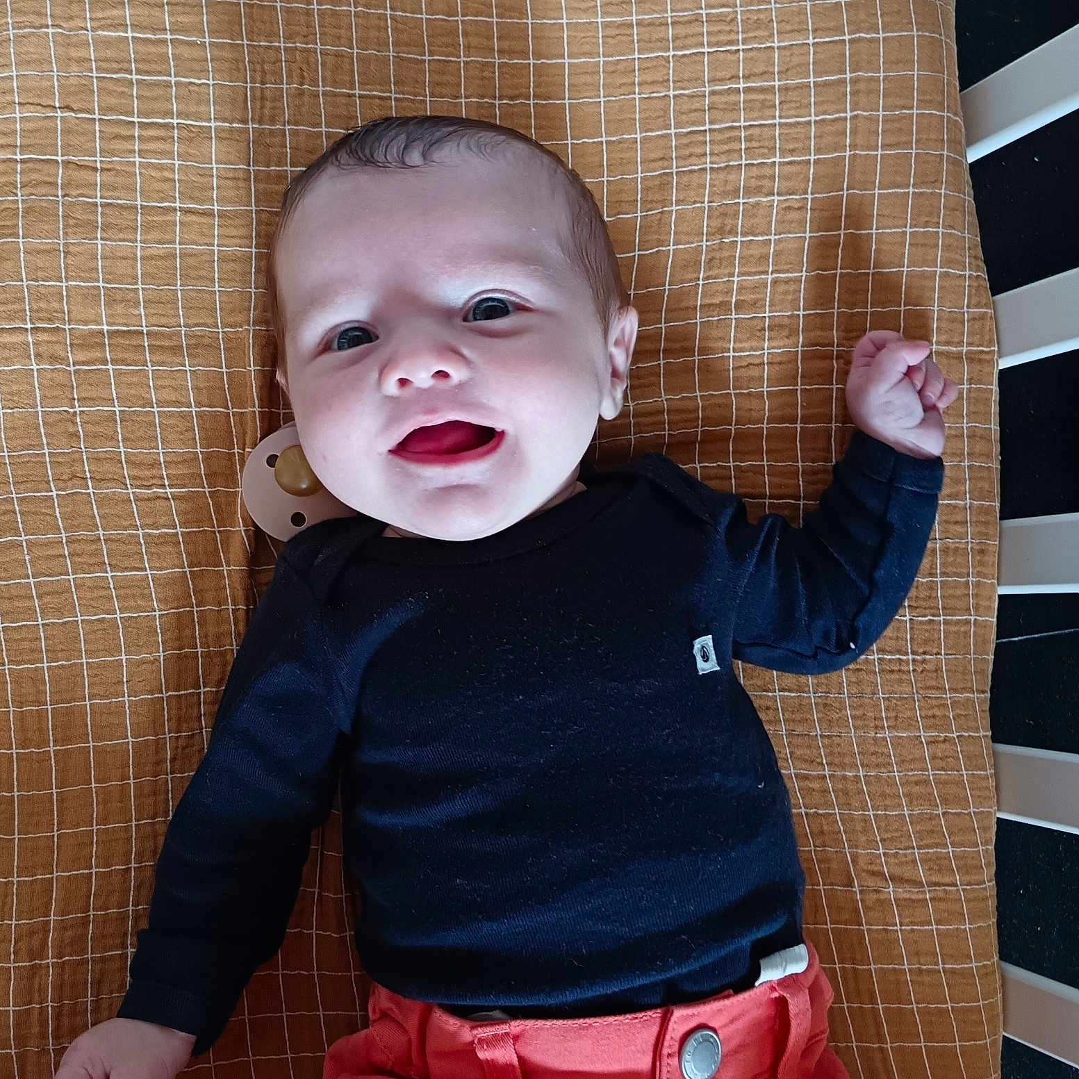 Céleste participe au concours pour gagner de l'argent avec cette photo : baby, baby_gear, bed, black_shirt, blanket, child, clothing, coral_pants, crib, cute, face, hands, happy, head, indoors, infant, lying_down, pacifier, person, smiling