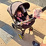 baby, sunglasses, stroller, outdoor, sidewalk, sunlight, shadow, person, hand, tattoo, footwear, casual_clothing, infant, plastic, wheel, canopy, safety_harness, toy, concrete, urban