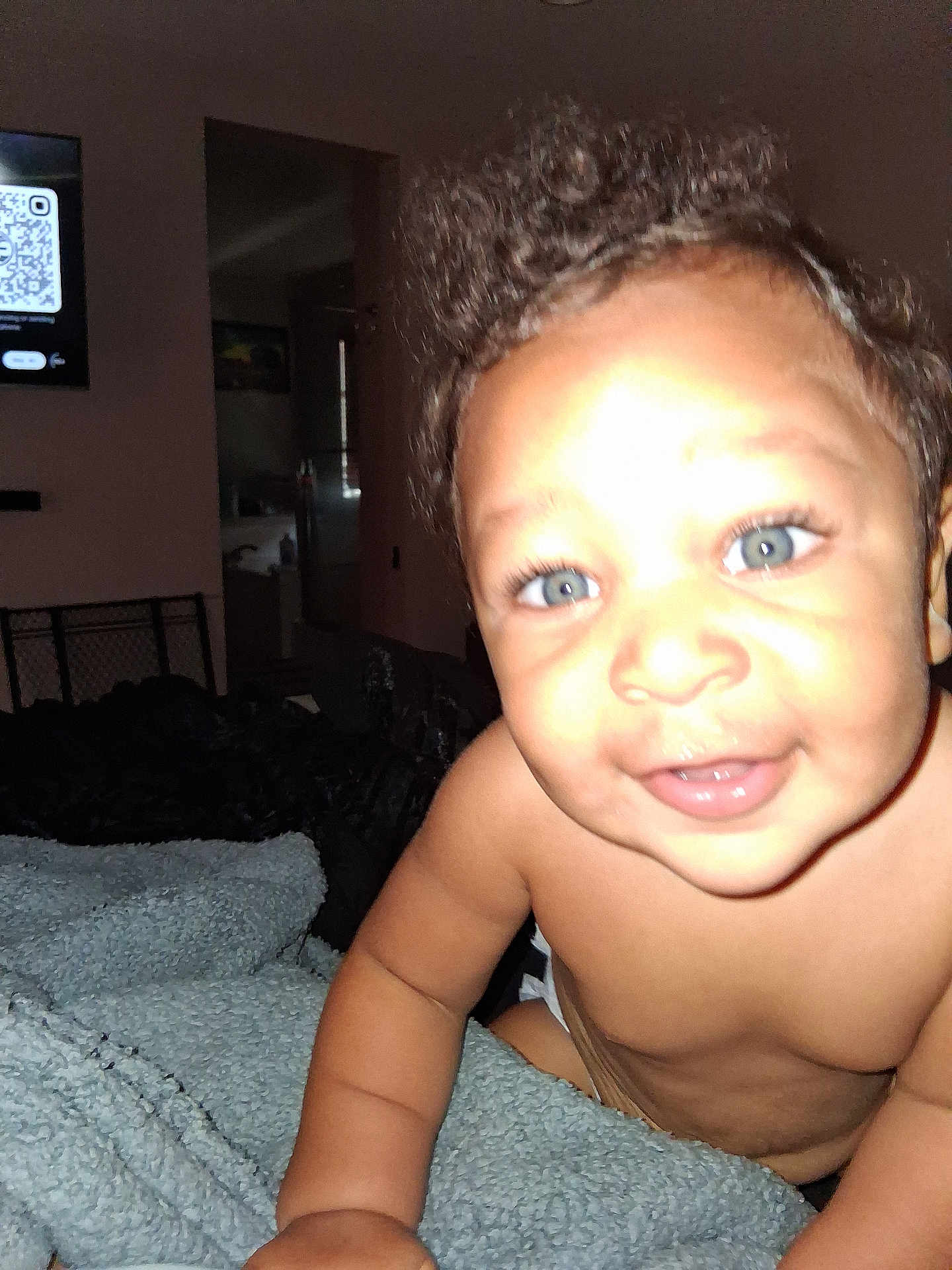 Saleem is registered to the contest to win money with this photo: baby, child, smiling, blue_eyes, curly_hair, blanket, indoor, close_up, face, skin, arm, diaper, furniture, television, qr_code, happy, cute, infant, person, portrait