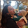 woman, baby, smile, laugh, outdoor, cafe, carousel, hat, jacket, blanket, hand, ring, tree, building, light, glass, drink, joy, happy, family