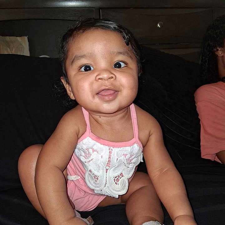 Emma Raquel Ward is registered to the contest to win money with this photo: baby, black_background, child, curly_hair, cute, face, footwear, furniture, hand, happy, indoors, infant, person, pink_outfit, sandals, sitting, skin, smiling, toes, young_child