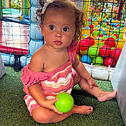 Zhavia is registered to the contest to win money with this photo: toddler, child, curly_hair, pink_dress, green_ball, play_area, ball_pit, netting, artificial_grass, indoor_playground, colorful, toy, cute, sitting, young_child, hair_clip, expression, daylight, play, fun