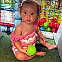 toddler, child, curly_hair, pink_dress, green_ball, play_area, ball_pit, netting, artificial_grass, indoor_playground, colorful, toy, cute, sitting, young_child, hair_clip, expression, daylight, play, fun
