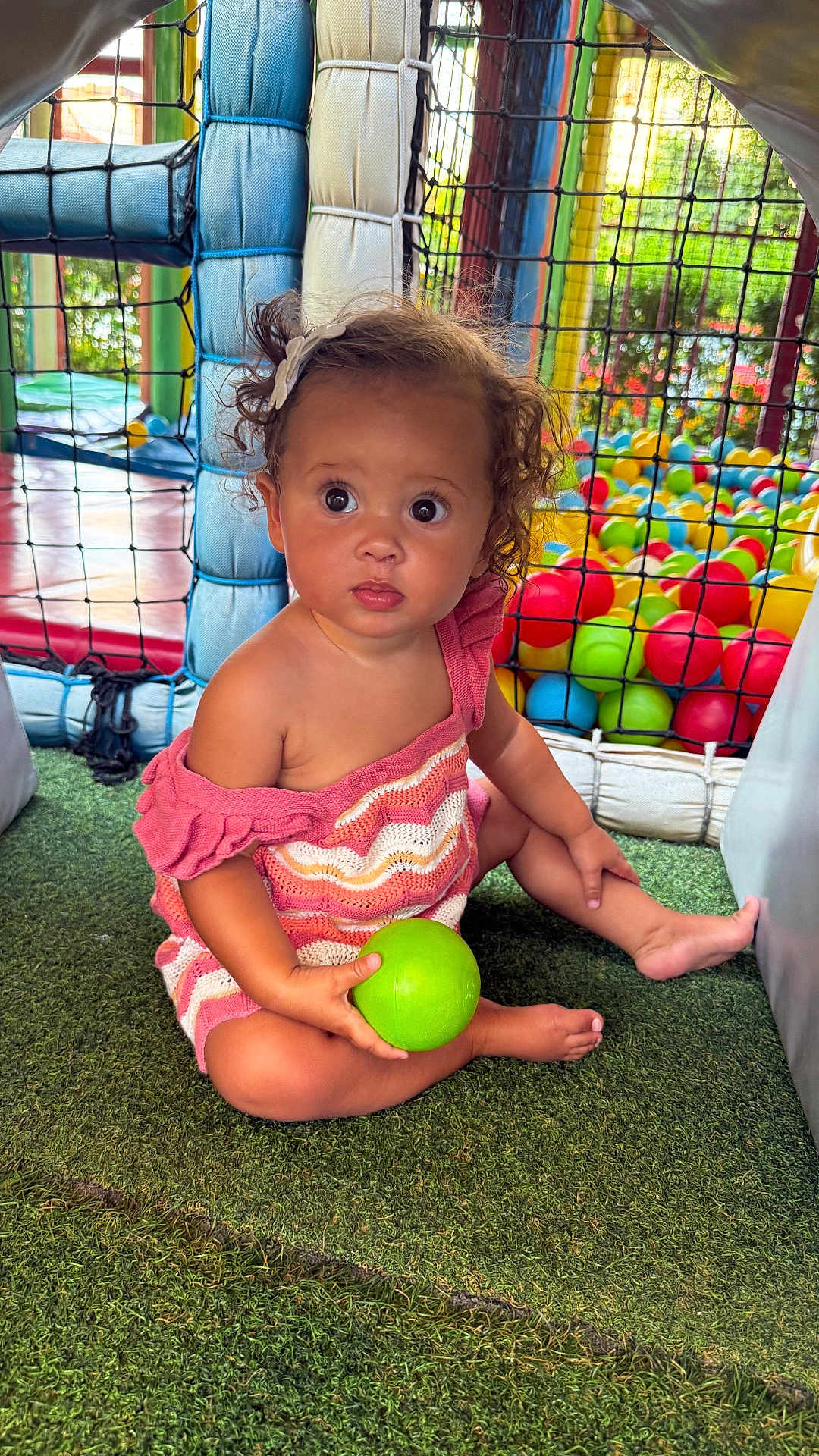 Zhavia is registered to the contest to win money with this photo: toddler, child, curly_hair, pink_dress, green_ball, play_area, ball_pit, netting, artificial_grass, indoor_playground, colorful, toy, cute, sitting, young_child, hair_clip, expression, daylight, play, fun