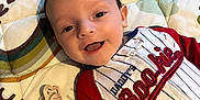 Easton Jo joined the competition — help win amazing prizes! baby, baby_boy, child, clothing, cute, face, happy, head, indoors, infant, lying_down, onesie, pacifier, person, portrait, quilt, red_sleeves, smiling, text_on_clothing, white_stripes