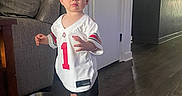 Sawyer is registered to the contest to win money with this photo: blond_hair, boy, child, couch, doorway, football_jersey, home, indoors, kitchen, living_room, natural_light, pet_bowl, portrait, serious_expression, sneakers, sportswear, standing, toddler, window, wood_floor