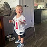 blond_hair, boy, child, couch, doorway, football_jersey, home, indoors, kitchen, living_room, natural_light, pet_bowl, portrait, serious_expression, sneakers, sportswear, standing, toddler, window, wood_floor