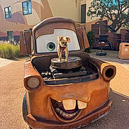 Squirt is registered to the contest to win money with this photo: animal, building, cartoon_car, curious, dog, engine, eyes, face, grass, mural, outdoor, pet, playful, rusty, sculpture, shadow, sidewalk, small_dog, sunlight, teeth
