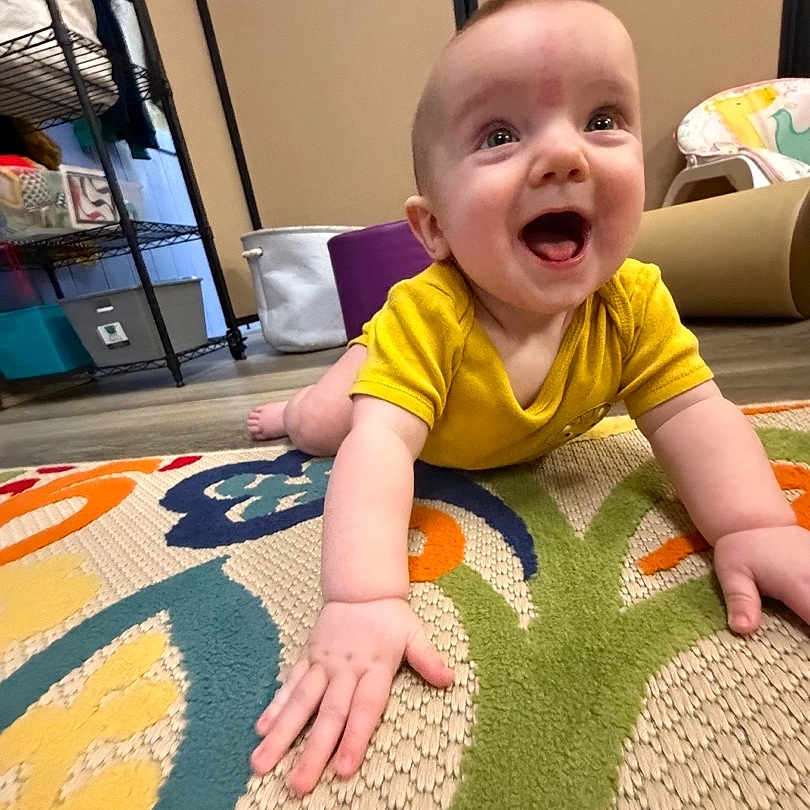 Vernon is registered to the contest to win money with this photo: baby, child, colorful, crawling, cute, expression, floor, furniture, hand, happy, head, home, indoor, infant, person, rug, smiling, storage_shelf, toy, yellow_shirt