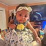 baby, child, indoor, dress, headband, flower, curly_hair, person, smiling, holding, furniture, television, bottle, room, hand, clothing, face, cute, infant, portrait