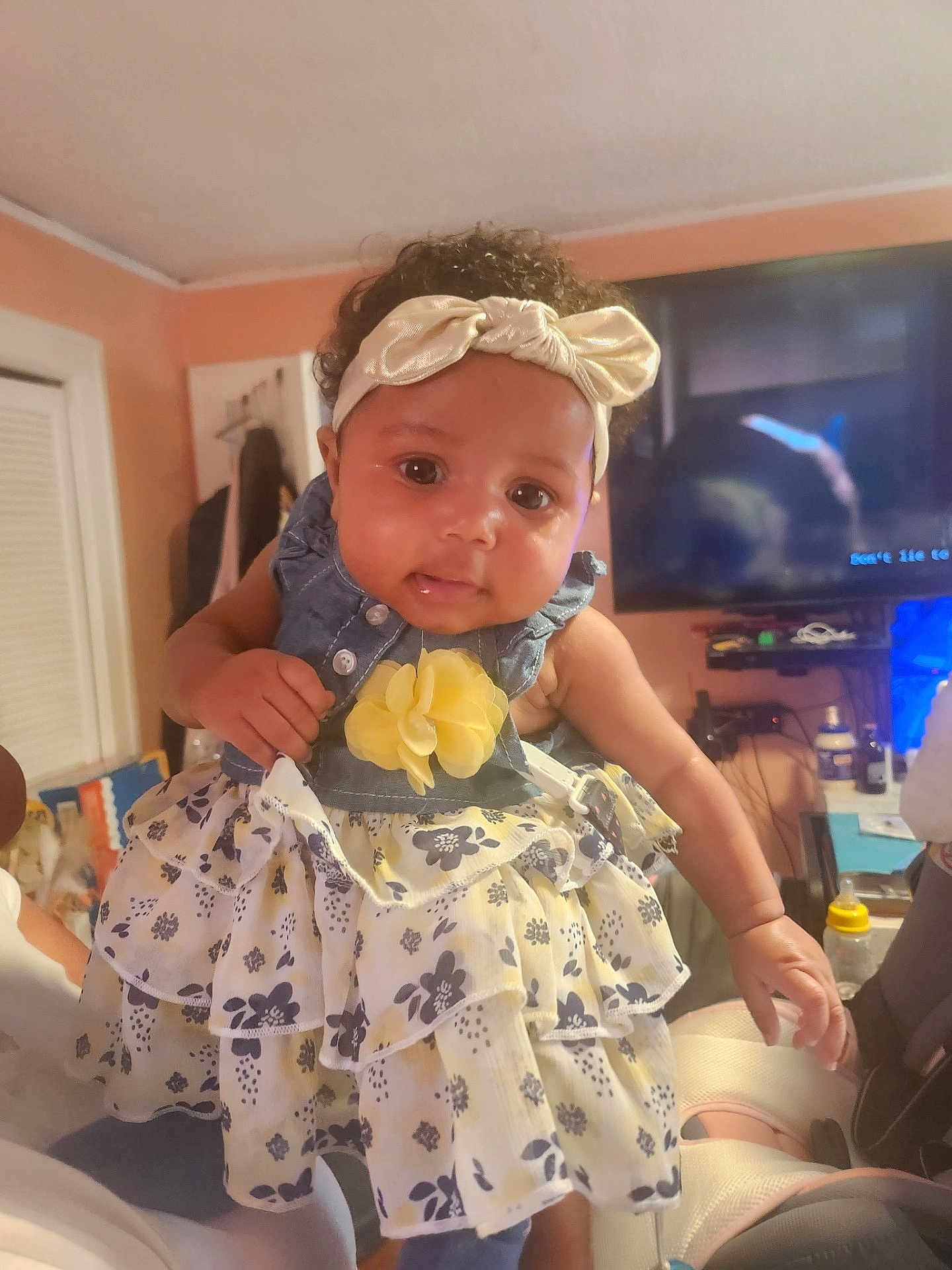 Ta'Lanni joined the competition — help win amazing prizes! baby, child, indoor, dress, headband, flower, curly_hair, person, smiling, holding, furniture, television, bottle, room, hand, clothing, face, cute, infant, portrait