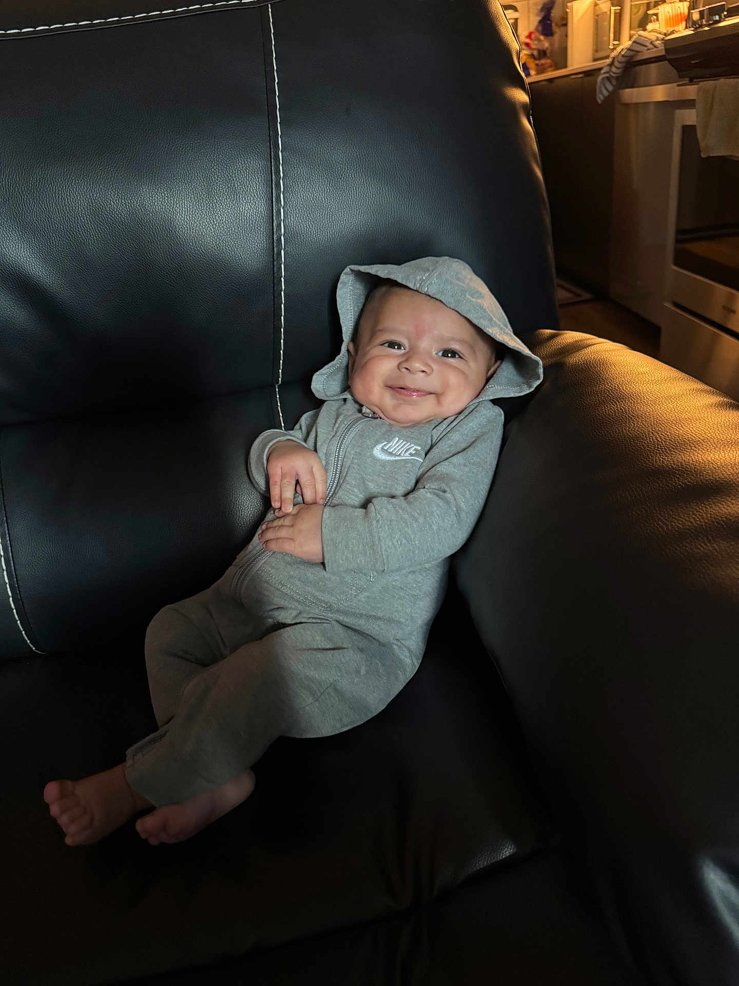 Jeremiah joined the competition — help win amazing prizes! baby, smiling, hoodie, gray_clothing, nike, infant, leather_couch, indoor, cozy, happy, child, person, casual, relaxed, comfort, living_room, candid, portrait, small_hands, barefoot