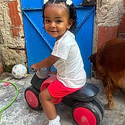 Kieza a rejoint le concours — aidez-le/la à gagner de superbes lots ! toddler, child, toy_vehicle, red, black, white_shirt, red_shorts, white_socks, white_shoes, blue_door, dog, concrete_floor, outdoor, hair_bows, curly_hair, play, pet, smile, fence, wall