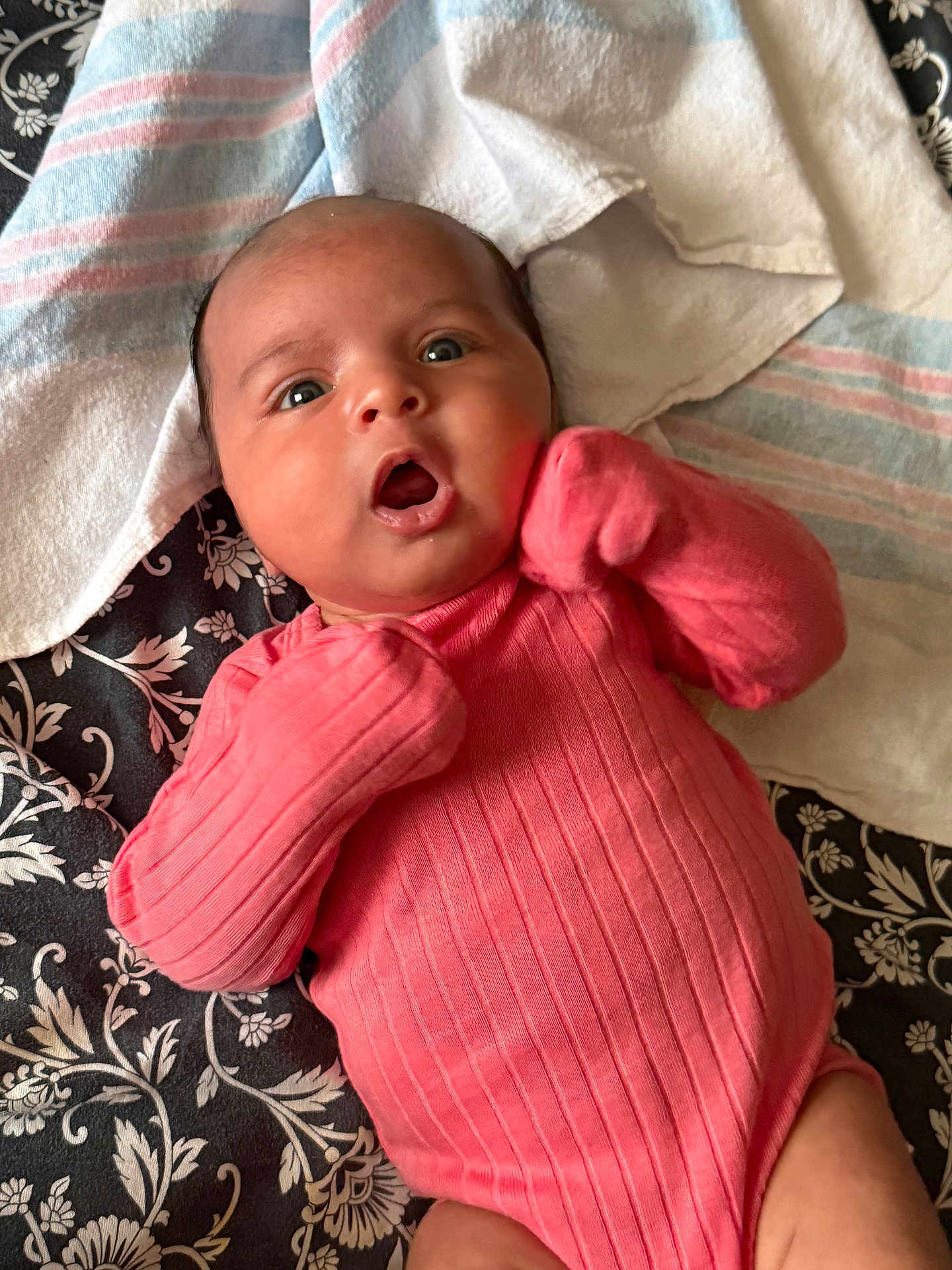 Carol is registered to the contest to win money with this photo: apparel, baby, blanket, child, cute, expression, face, hands, head, indoor, infant, lying_down, newborn, onesie, pattern, person, pink_clothing, portrait, skin, towel