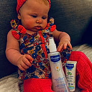 Lya a rejoint le concours — aidez-le/la à gagner de superbes lots ! baby, infant, child, floral_dress, red_headband, red_leggings, mustela_products, bottle, tube, sofa, blanket, indoor, curious, sitting, skin_care, person, face, hand, cozy, cute