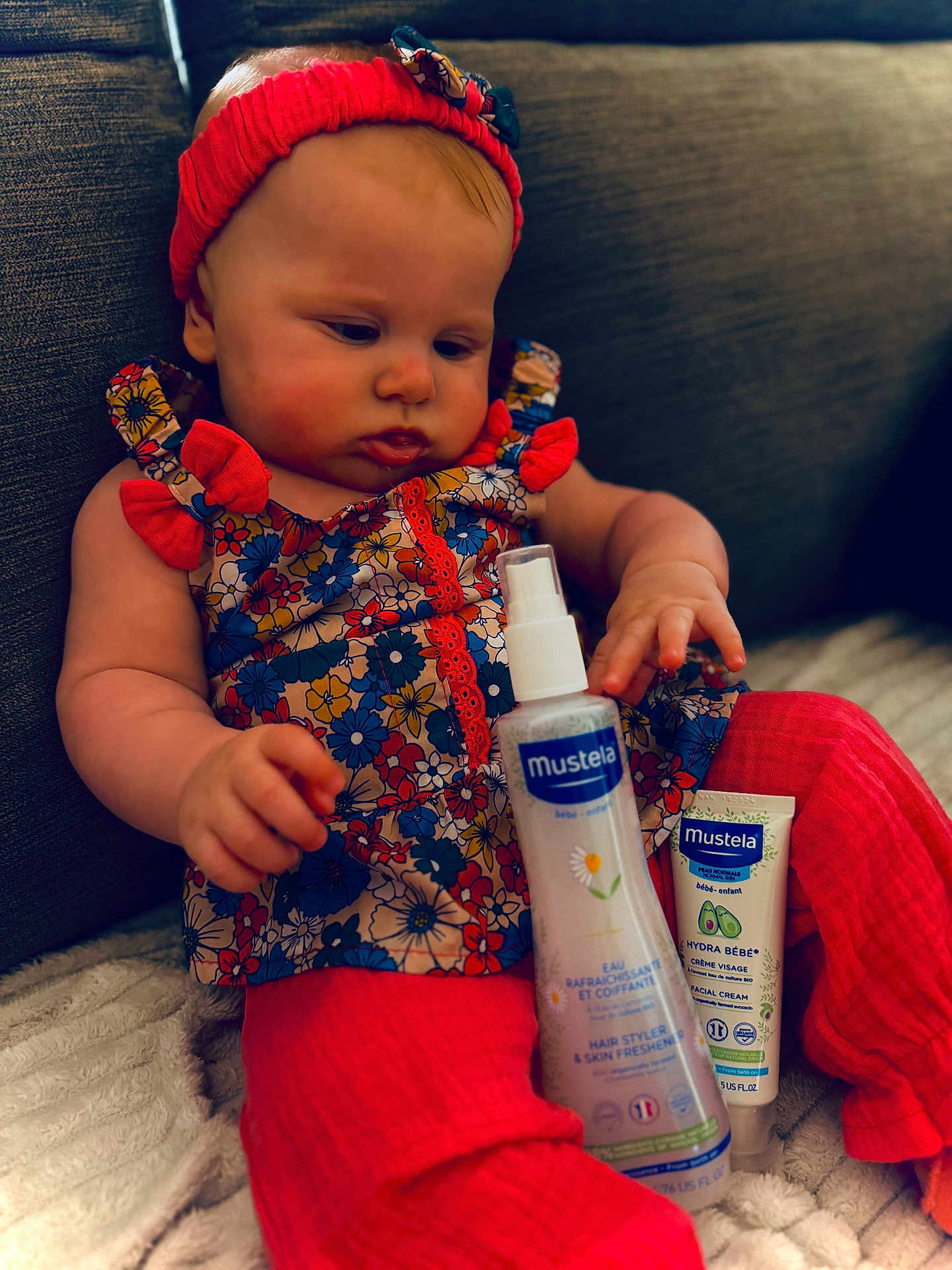 Lya a rejoint le concours — aidez-le/la à gagner de superbes lots ! baby, infant, child, floral_dress, red_headband, red_leggings, mustela_products, bottle, tube, sofa, blanket, indoor, curious, sitting, skin_care, person, face, hand, cozy, cute