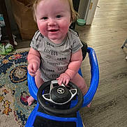 Giovanni is registered to the contest to win money with this photo: baby, child, smiling, toy_car, indoor, floor, rug, steering_wheel, blue, clothing, person, happy, sitting, wooden_floor, hand, face, cheeks, hair, play, home