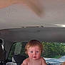 baby, blonde_hair, blue_eyes, car_headrest, car_seat, car_window, child, child_safety, curious, curly_hair, infant, outdoor, person, reaching_hand, rear_view, road, seatbelt, sunlight, toddler, vehicle_interior
