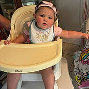 Clara is registered to the contest to win money with this photo: baby, high_chair, bib, pink_headband, child, indoor, curious, rosy_cheeks, tableware, china_cabinet, blanket, hello_kitty, tile_floor, person, cute, infant, furniture, reaching_out, pink_clothing, foot