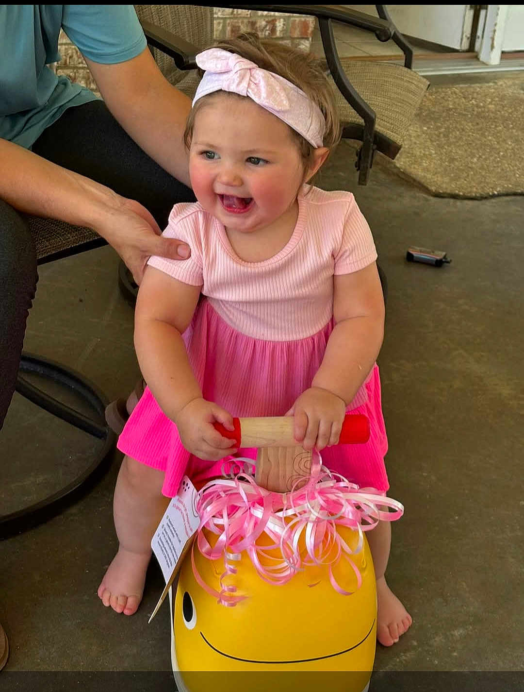 Clara joined the competition — help win amazing prizes! toddler, child, pink_dress, headband, toy, yellow_toy, ribbons, smiling, happy, barefoot, adult_hand, outdoor, patio, concrete_floor, chair, person, supporting, cute, playtime, baby