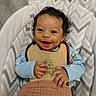 baby, smiling, infant, bib, blanket, cushion, chevron_pattern, clothing, curly_hair, happy, indoor, person, portrait, seat, soft_texture, young_child, cute, baby_girl, baby_boy, comfortable
