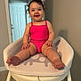 baby, child, high_chair, pink_clothing, smiling, indoor, person, sitting, curly_hair, happy, furniture, door, wall, floor, home, infant, cute, young_child, plush_seat, legs
