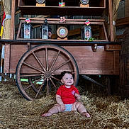 Manuela joined the competition — help win amazing prizes! baby, child, hay, wooden_cart, wheel, lantern, red_shirt, smile, sitting, indoor, rustic, flag, decor, cute, happy, barn, person, infant, floor, wood