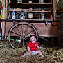 baby, child, hay, wooden_cart, wheel, lantern, red_shirt, smile, sitting, indoor, rustic, flag, decor, cute, happy, barn, person, infant, floor, wood