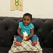 Priscillya a rejoint le concours — aidez-le/la à gagner de superbes lots ! baby, child, couch, cushions, toy, milestone_card, blue_clothing, smile, indoor, wall, patterned_cushions, happy, sitting, infant, feet, face, person, home, decor, cute