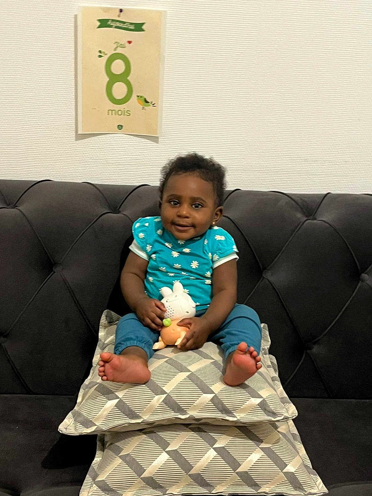 Priscillya a rejoint le concours — aidez-le/la à gagner de superbes lots ! baby, child, couch, cushions, toy, milestone_card, blue_clothing, smile, indoor, wall, patterned_cushions, happy, sitting, infant, feet, face, person, home, decor, cute