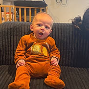 Karter participe au concours pour gagner de l'argent avec cette photo : baby, infant, orange_onesie, couch, indoor, expression, mouth_open, furniture, baby_gate, home, child, person, seated, ribbed_texture, cozy_clothing, curly_hair, wooden_furniture, background, domestic, happy