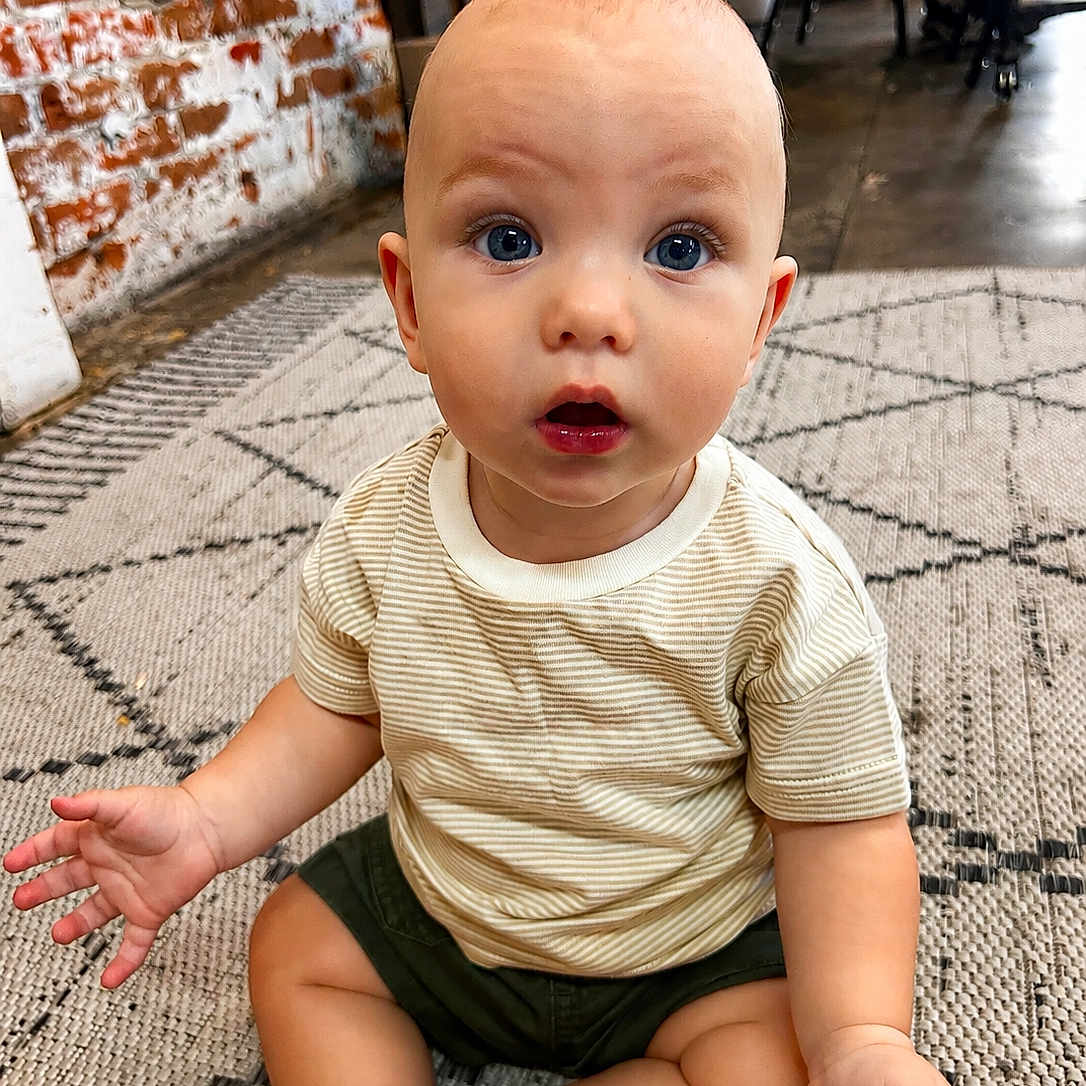 Bentlee Barron is registered to the contest to win money with this photo: baby, barefoot, blue_eyes, brick_wall, casual, child, clothing, curious, expression, face, floor, furniture, home, indoor, patterned_rug, person, rug, shorts, sitting, striped_shirt