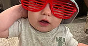 Bentlee Barron joined the competition — help win amazing prizes! baby, red_glasses, bucket_hat, toddler, indoor, carpet, tattoo, adult, phone, couch, shirt, cute, child, hand, sitting, face, glasses, person, home, casual