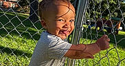 Elijah is registered to the contest to win money with this photo: toddler, child, smiling, outdoor, fence, chain_link_fence, sunlight, backyard, grass, trees, house, portrait, standing, playing, happy, shorts, t_shirt, face, summer, daylight