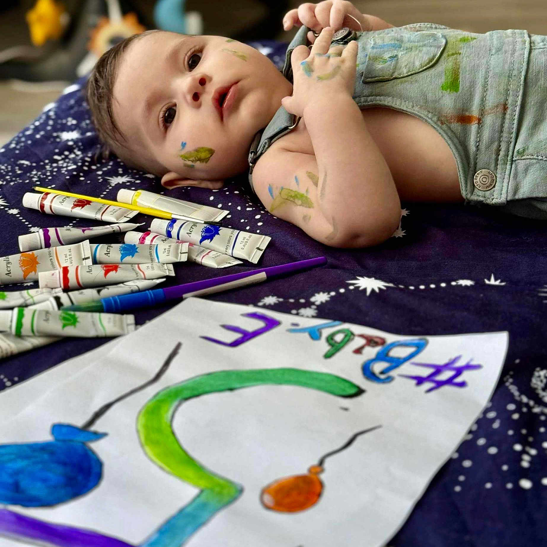 Elis participe au concours pour gagner de l'argent avec cette photo : artwork, baby, blanket, child, closeup, colorful, creativity, drawing, expression, face, hand, indoor, laying_down, messy, overalls, paint, paint_brush, paint_tubes, paper, toddler