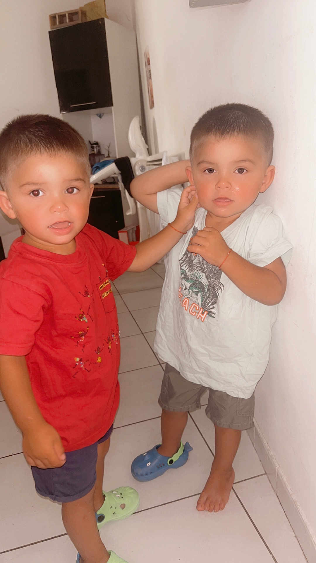 Kayden Aaron participe au concours pour gagner de l'argent avec cette photo : child, boy, indoor, tiled_floor, red_shirt, white_shirt, shorts, slippers, barefoot, wall, kitchen, cabinet, floor, furniture, person, tile, standing, casual_clothing, home, siblings