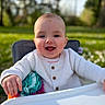 baby, smiling, high_chair, white_sweater, outdoor, daylight, colorful_toy, happy, child, portrait, person, greenery, nature, grass, blurred_background, cute, infant, face, seated, warm_clothing