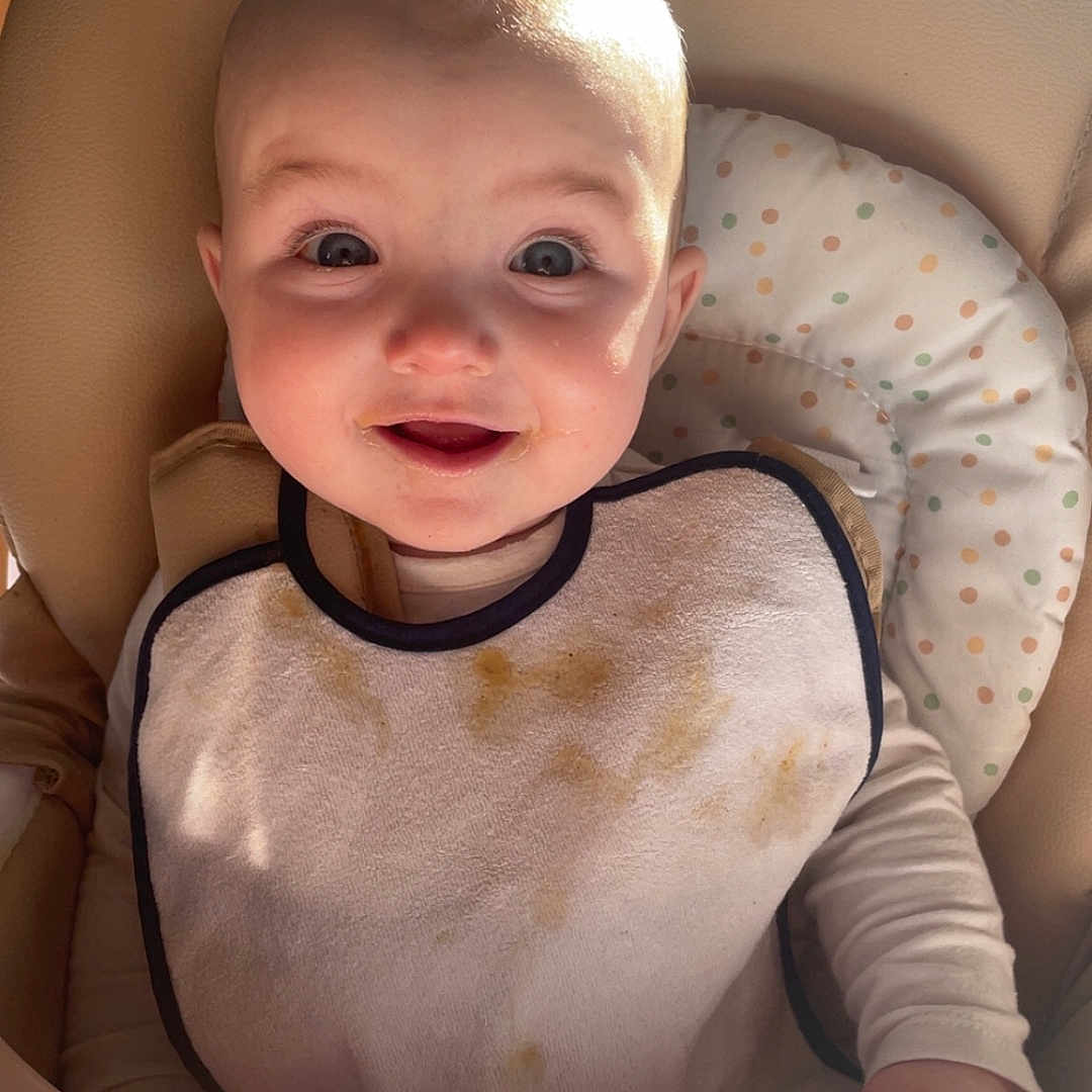 Inaya a rejoint le concours — aidez-le/la à gagner de superbes lots ! baby, bib, high_chair, smiling, messy, indoor, child, face, seat, feeding, happy, young_child, infant, cute, person, clothing, head, light, sunlight, expression