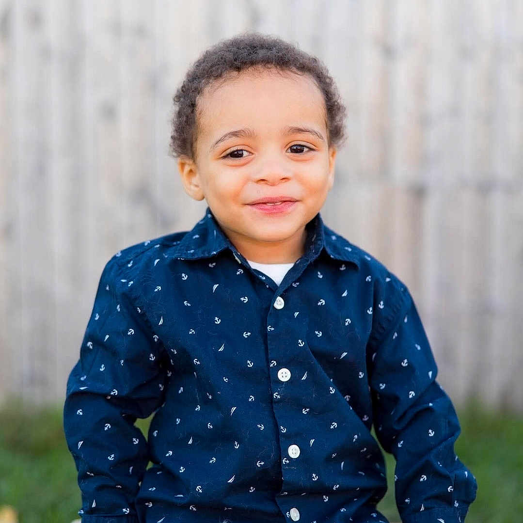 Izaiah is registered to the contest to win money with this photo: toddler, child, smiling, portrait, outdoor, blue_shirt, patterned_clothing, curly_hair, happy, sitting, wooden_fence, grass, casual, buttoned_shirt, cute, face, young_child, person, daylight, nature