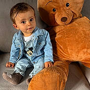 Julien a rejoint le concours — aidez-le/la à gagner de superbes lots ! toddler, child, denim_jacket, denim_overalls, socks, plush_toy, teddy_bear, couch, indoor, cute, smiling, face, person, seated, soft_toy, home, casual_clothing, brown, gray, fabric